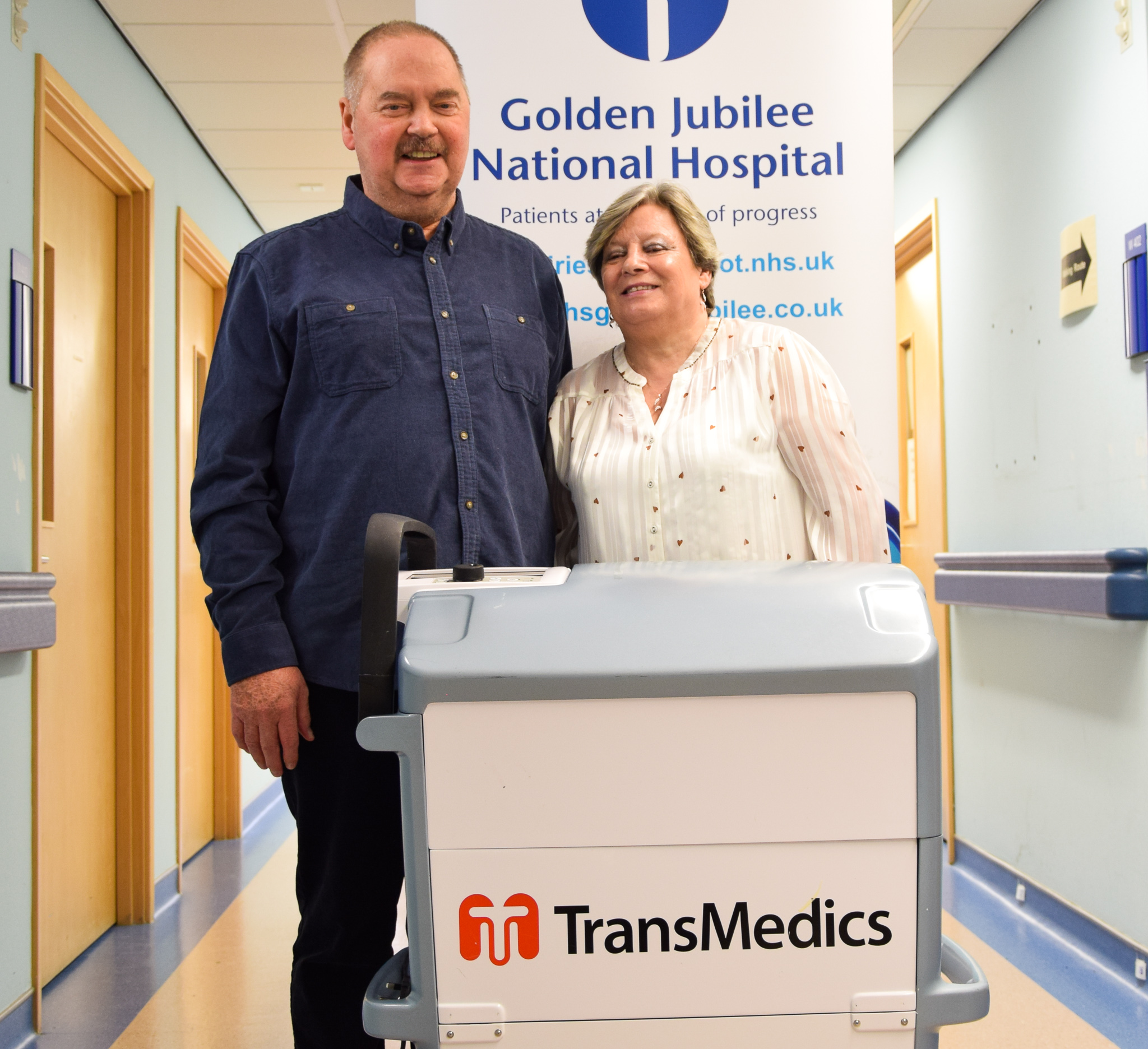 Colin Davidson and wife, Susan, smiling in front of NHS Golden Jubilee banner.