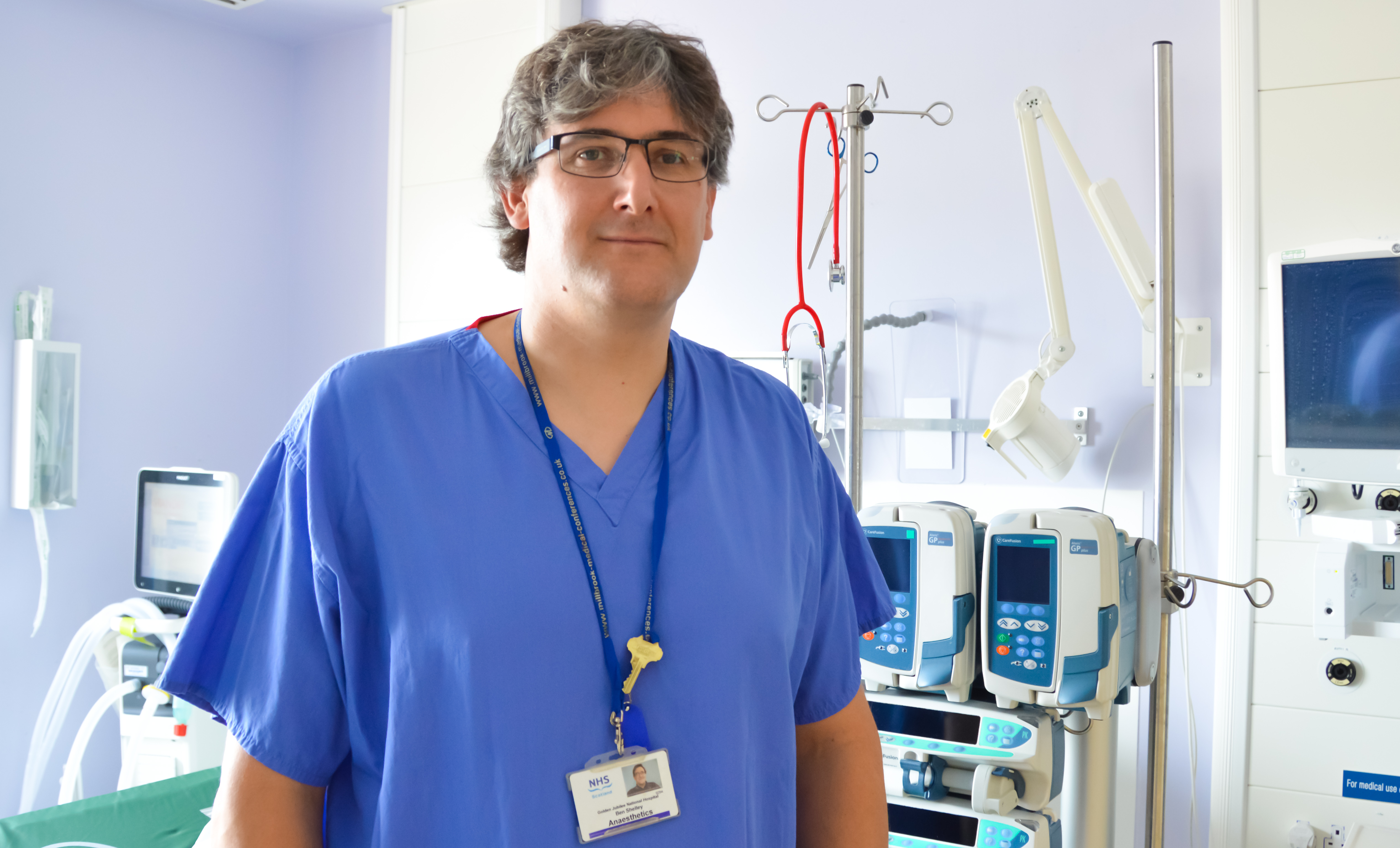 Ben Shelley in NHS uniform, standing in front of clinical machines.